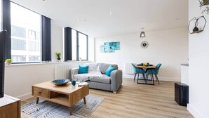 A modern living room with a grey sofa, a wooden coffee table, and a dining area with blue chairs. - Luxurious Stylish Apartment (Stretford)