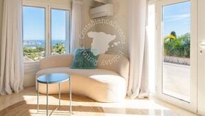 Un saló modern amb un sofà corbat, una taula rodona petita i vista al mar a través de grans finestres.