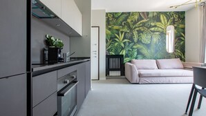 A modern kitchen with grey cabinets and stainless steel appliances, a dining area with a white sofa and a black chair, and a large tropical leaf-patterned wall mural. - Daplace - Vico Apartment (Milano)