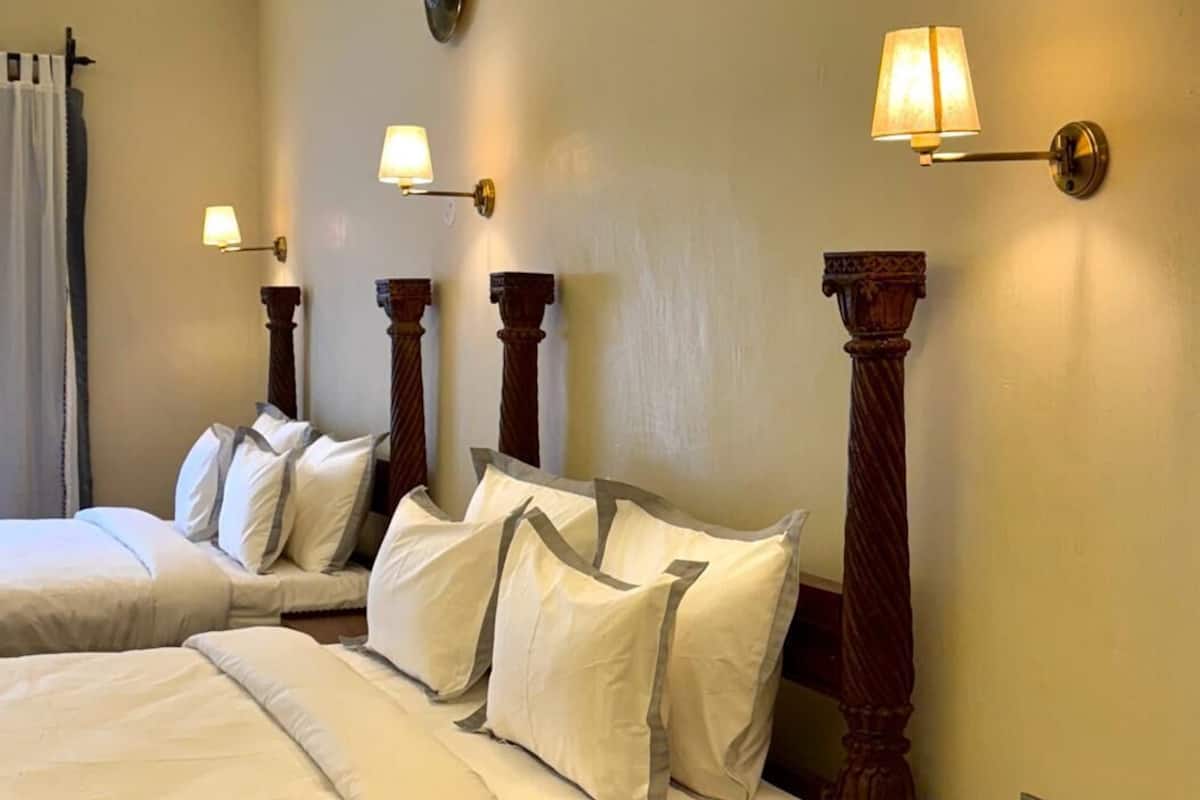 A hotel room with two beds, each with white linens and multiple pillows. There are wall-mounted lamps on either side of the beds.