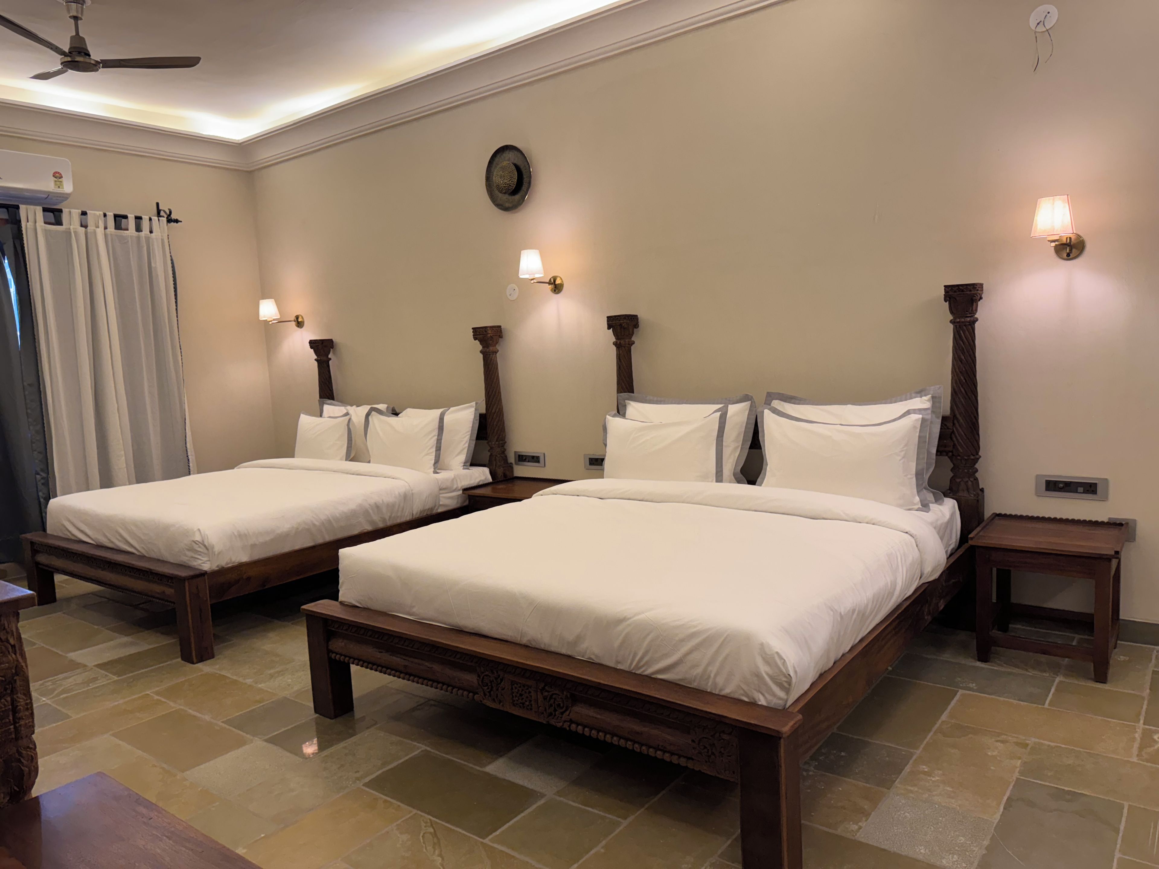 A hotel room with two wooden beds, white linens, a ceiling fan, and wall-mounted lamps.
