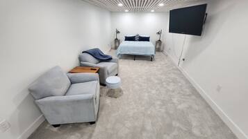 Une pièce moderne et bien éclairée, avec un canapé gris, un petit pouf et un lit recouvert d’une couverture bleue.