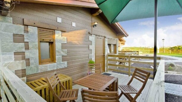 Um deck de madeira com mesa e cadeiras, um muro de pedra e um guarda-sol verde.