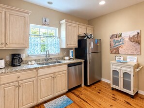A kitchen with wooden cabinets, a stainless steel refrigerator, a coffee maker, and a window with a view of greenery. - Whispering Pines Cottage–Peaceful Fort Morgan Retreat | Pool | Close to Beach (Gulf Shores)