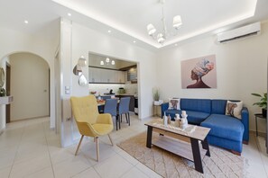 Ein modernes Wohnzimmer mit einer Couch, einem Couchtisch und einem gelben Sessel. Im Hintergrund ist eine Küche mit einem Essbereich zu sehen.