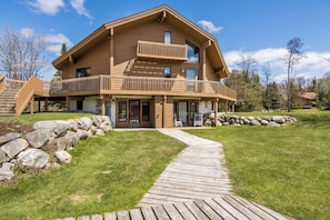 A two-story log cabin with a balcony, a stone pathway, and a stone wall. - Greens & Glides Getaway at Schuss Mountain (Mancelona)