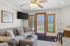 A living room with a sofa, a ceiling fan, a TV mounted on the wall, and a sliding glass door leading to an outdoor area. - Greens & Glides Getaway at Schuss Mountain (Mancelona)