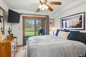 Ein Schlafzimmer mit Bett, Deckenventilator, Fernseher, Schreibtisch, Stuhl und einer Tür, die auf einen Balkon führt.