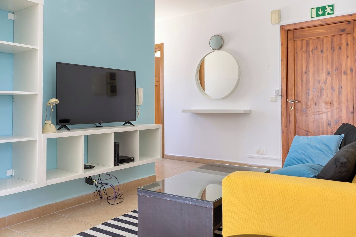 A modern living room with a flat-screen TV on a white stand, a yellow sofa, and a wooden door with an emergency exit sign.