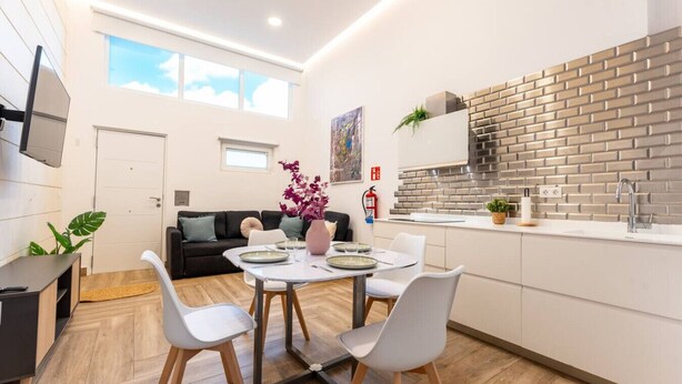 Eine moderne Küche mit Ziegelrückwand, ein Essbereich mit Tisch und Stühlen sowie ein Wohnbereich mit Sofa und Fernseher.