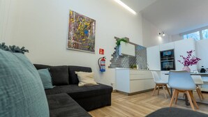 Ein modernes Wohnzimmer mit einer schwarzen Couch, einer kleinen Küchenzeile mit weißer Arbeitsplatte und einem Esstisch mit Stühlen.