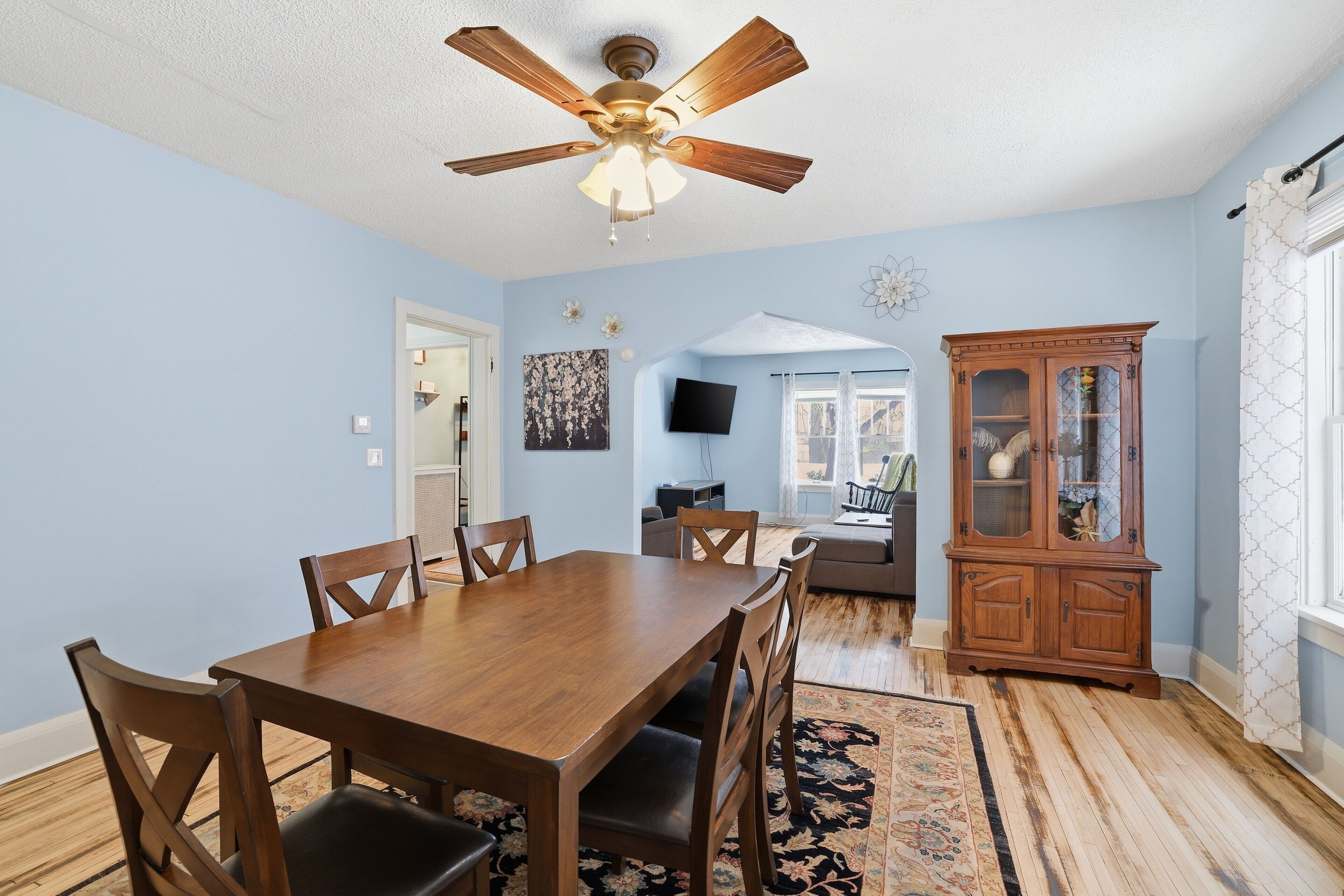 A dining room with a wooden table and chairs, a ceiling fan, and a patterned rug.