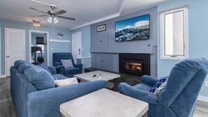 A modern living room with a fireplace, blue sofas, a ceiling fan, and a large TV on the wall. - Bridges 76 2 Bedroom Condo (Warren)