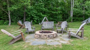 A stone fire pit surrounded by Adirondack chairs in a wooded backyard. - Bridges 76 2 Bedroom Condo (Warren)