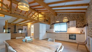 A spacious kitchen with a stone wall, wooden beams, and a large dining table with chairs. - The Stone House – naturaleza y confort (villacarriedo)