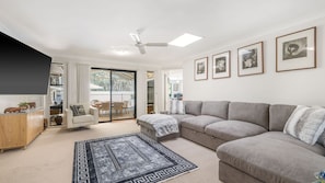 Una sala de estar moderna con un sofá modular gris, un sillón blanco, una mesa de centro y un tapete estampado. Hay cuadros en la pared y un ventilador de techo.