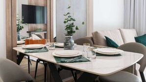 Een eetgedeelte met een witte tafel voor vier personen, omringd door stoelen. Op de tafel staat een vaas met groene bladeren, en er is een flatscreen televisie aan de muur achter de tafel bevestigd.