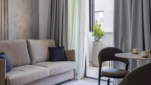 A modern living room with a sofa, a coffee table, and chairs. There is a large window with curtains, a potted plant, and a view of the outside. - Modern Studio Suite w/ Balcony & Queen Bed – Germasogeia Tourist Area, Limassol (Germasogeia)