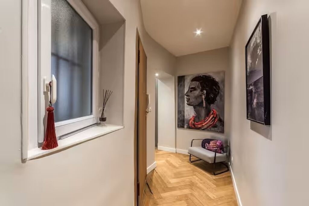 A hallway with a wooden floor, a window with a red vase, and a painting on the wall.