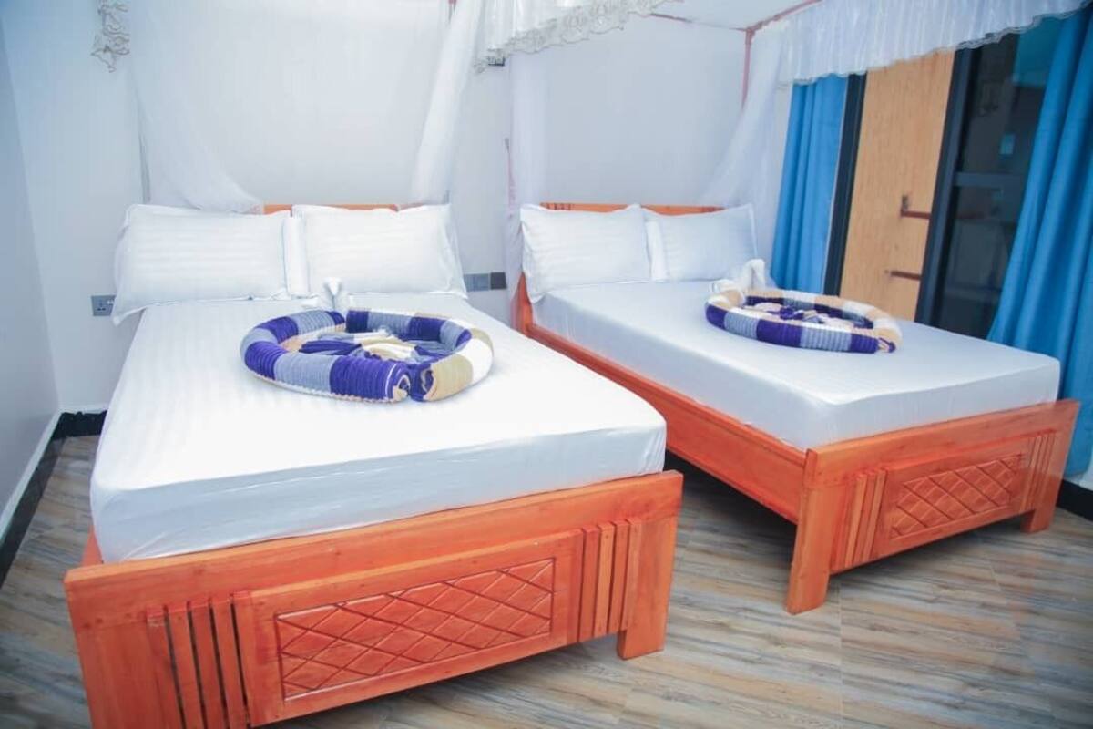 Dos camas individuales con estructuras de madera y ropa blanca, una mosquitera sobre las camas y una cortina azul en la ventana.