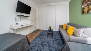 Een moderne woonkamer met een flatscreen televisie, een bank met gele kussens, een kleine tafel met een plant en een grijs vloerkleed.