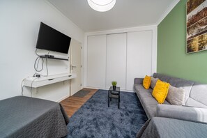 Ein modernes Wohnzimmer mit einem Flachbildfernseher, einem Sofa mit gelben Kissen, einem kleinen Tisch mit Pflanze und einem grauen Teppich.