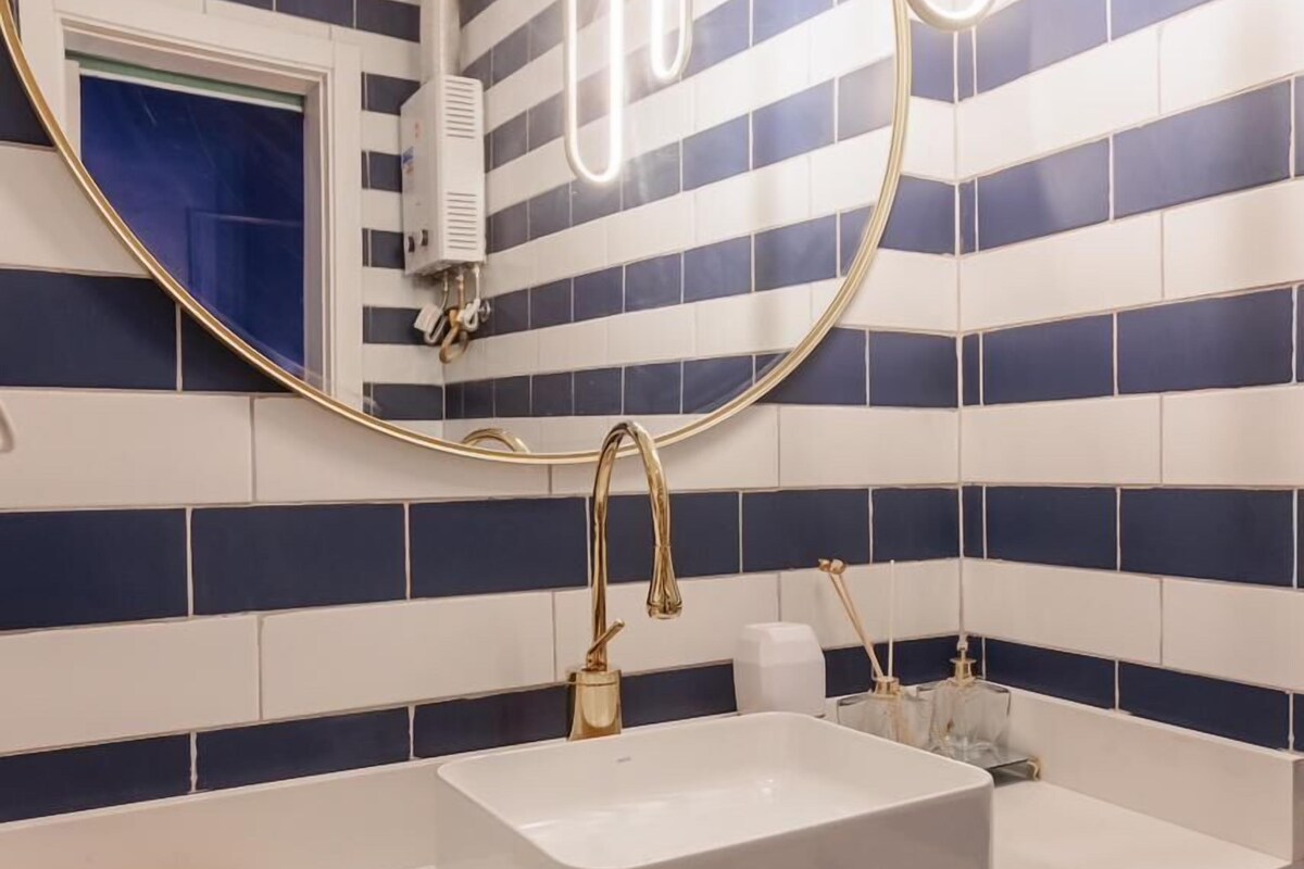 Un baño con lavamanos blanco, grifo dorado y un espejo grande con marco dorado. Las paredes están cubiertas de azulejos con un estampado azul y blanco a rayas.