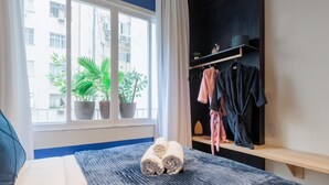 A bedroom with a bed, two potted plants, a window with a view of buildings, and a clothing rack with a robe and a towel. - StayHome Beira-Mar Copacabana Posto 6 (Rio de Janeiro)