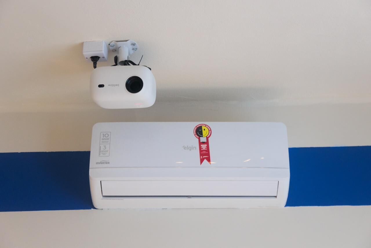 Un aire acondicionado fijado a la pared con un adhesivo de cinta roja que indica un premio y un proyector blanco instalado en el techo.