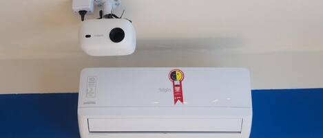 Un aire acondicionado fijado a la pared con un adhesivo de cinta roja que indica un premio y un proyector blanco instalado en el techo.