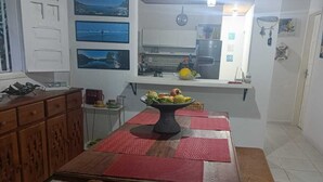 Un comedor con una mesa de madera, bancos y una silla de mimbre. Se ve una cocina al fondo con un frutero sobre la encimera.