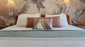 Quarto de hotel com uma cama grande, duas luminárias fixadas na parede e um mural de pássaros na parede.