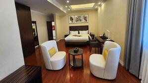 Habitación de hotel con cama, dos sillones blancos, una mesita redonda, suelo de madera e iluminación empotrada en el techo.