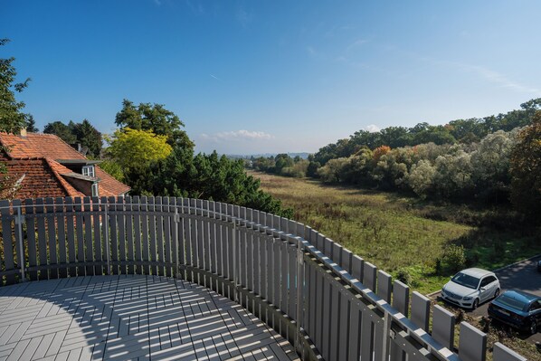Une vue depuis un balcon doté d’une balustrade en bois, donnant sur un champ verdoyant et des arbres, avec un bâtiment au toit rouge en arrière-plan.