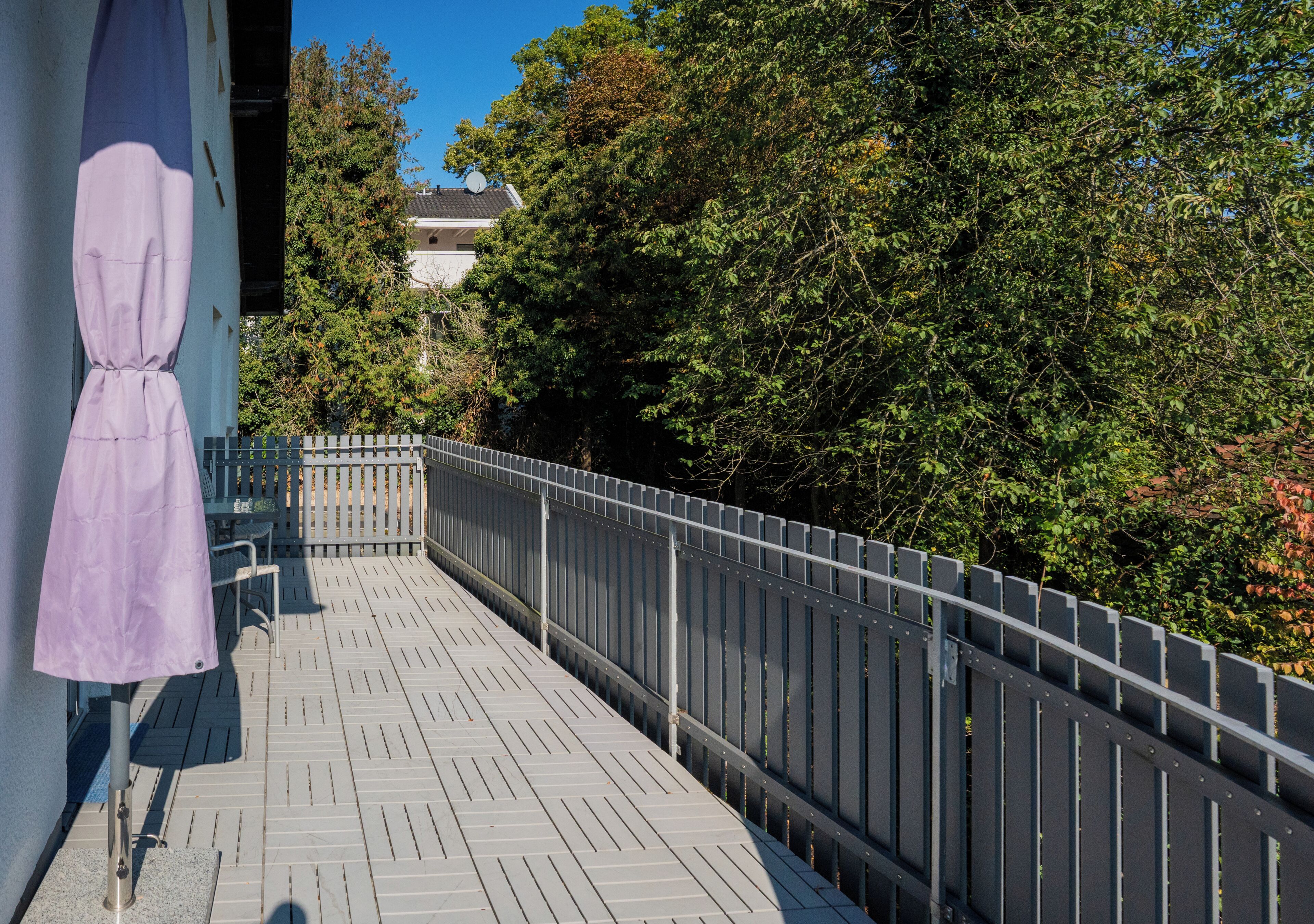 Una terraza de madera con barandal metálico, un área de estar cubierta y vistas a árboles y un edificio al fondo.