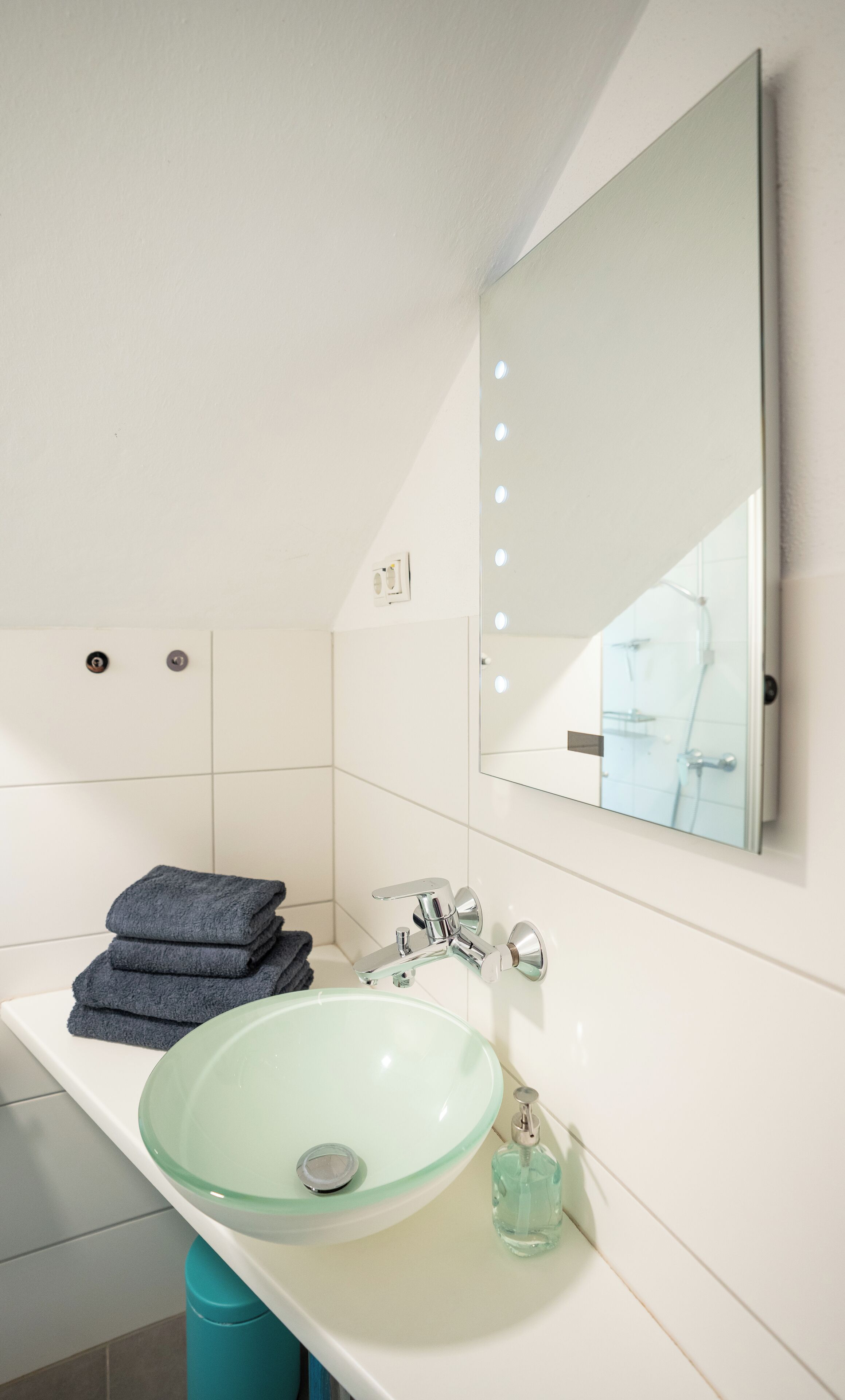 A bathroom with a glass sink, a mirror with lights, and a stack of towels.