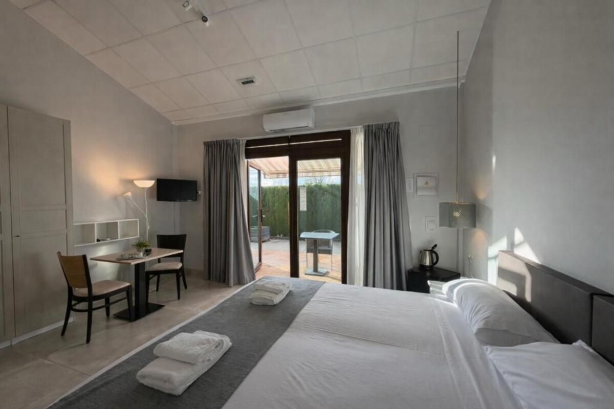 A hotel room with a bed, a desk, and a chair. There is a TV on the wall, a window with curtains, and a small table with a lamp.