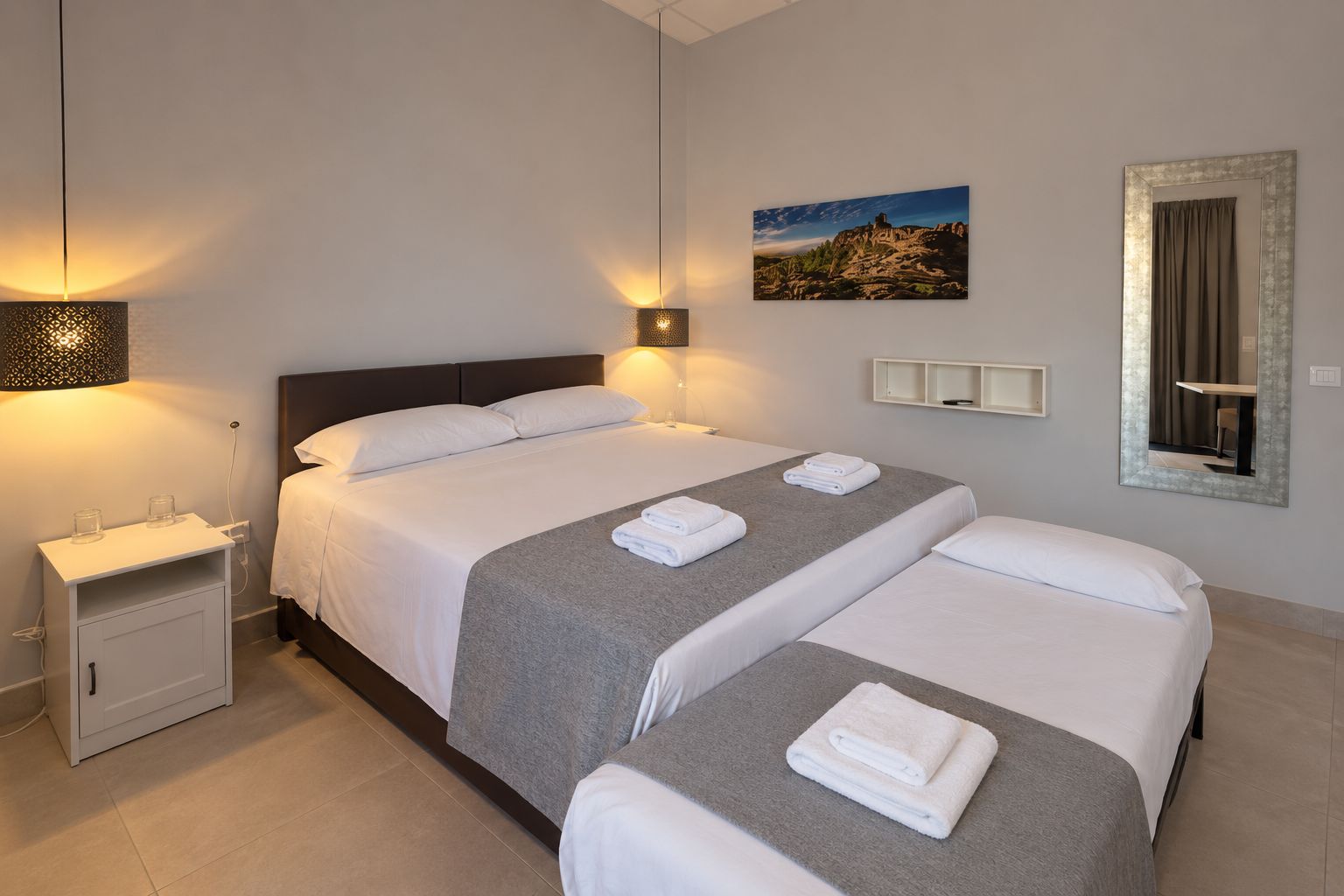 Habitación de hotel con dos camas, una mesita de noche, una lámpara de pared, un cuadro enmarcado y un espejo.