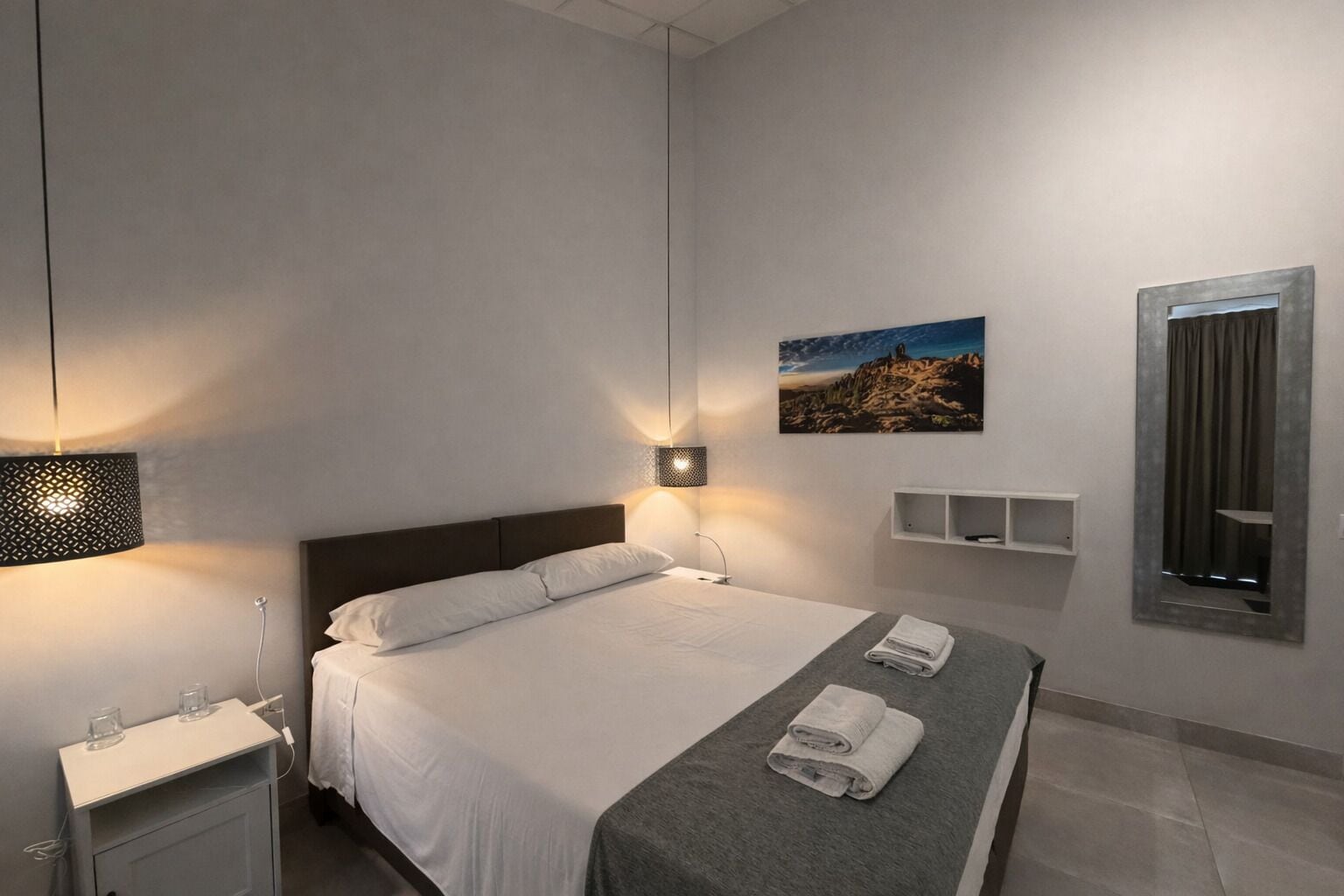 A modern hotel room with a neatly made bed, a bedside table with glasses, a wall-mounted lamp, a painting, and a mirror.