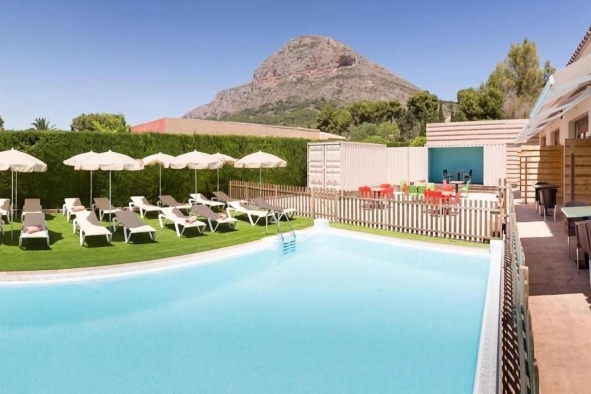 A swimming pool with lounge chairs and umbrellas, a wooden fence, and a mountain in the background.