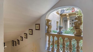 Un escalier orné de tableaux encadrés le long du mur, une balustrade surmontée de lions en pierre, et une vue sur un bâtiment visible à travers une arche.