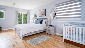 A bedroom with a bed, bedside tables, a crib, and a window with blinds. - Charming riverside cottage (Wasaga Beach)