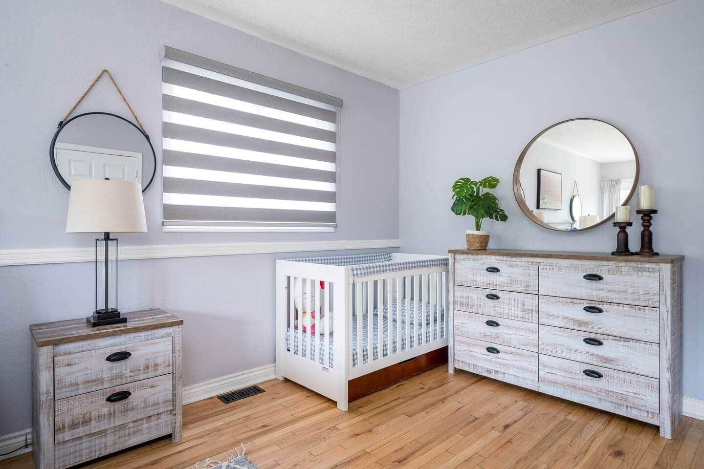 A room with a crib, a dresser with drawers, a lamp, a window with blinds, and a mirror on the wall.