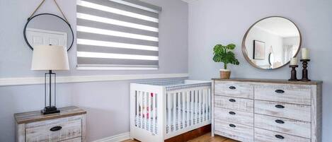 A room with a crib, a dresser with drawers, a lamp, a window with blinds, and a mirror on the wall.