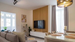 Uma sala de estar moderna com um sofá, uma mesa de jantar com cadeiras, uma TV na parede e uma janela com cortinas.