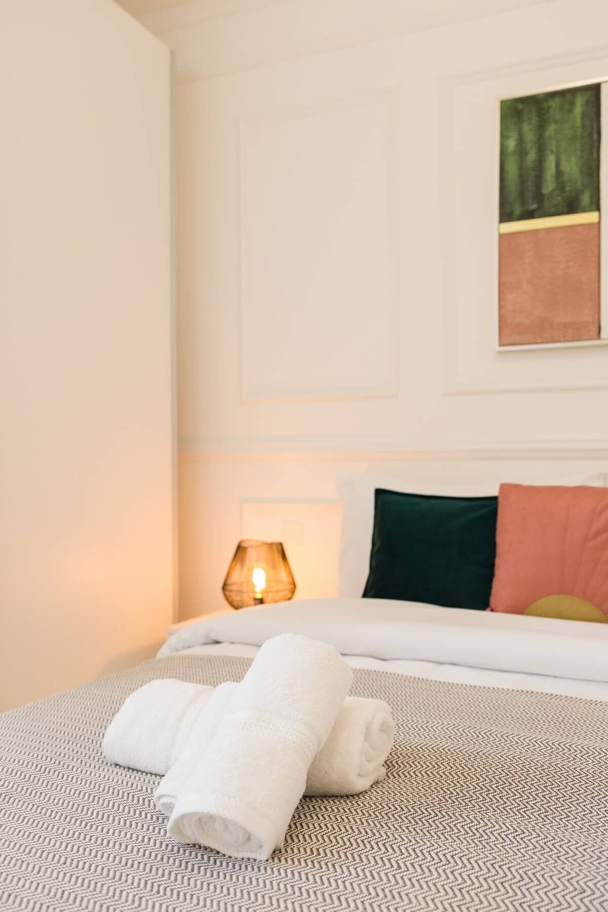 Un lit bien fait, avec deux serviettes enroulées, une lampe de chevet et un tableau accroché au mur.