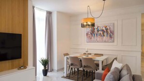 Uma sala de estar moderna com uma TV de tela plana, uma área de jantar com uma mesa e cadeiras brancas e um grande quadro abstrato na parede.
