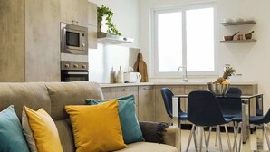 Een moderne woonkamer met een bank, een eethoek met stoelen, een keuken met apparatuur en een groot raam.