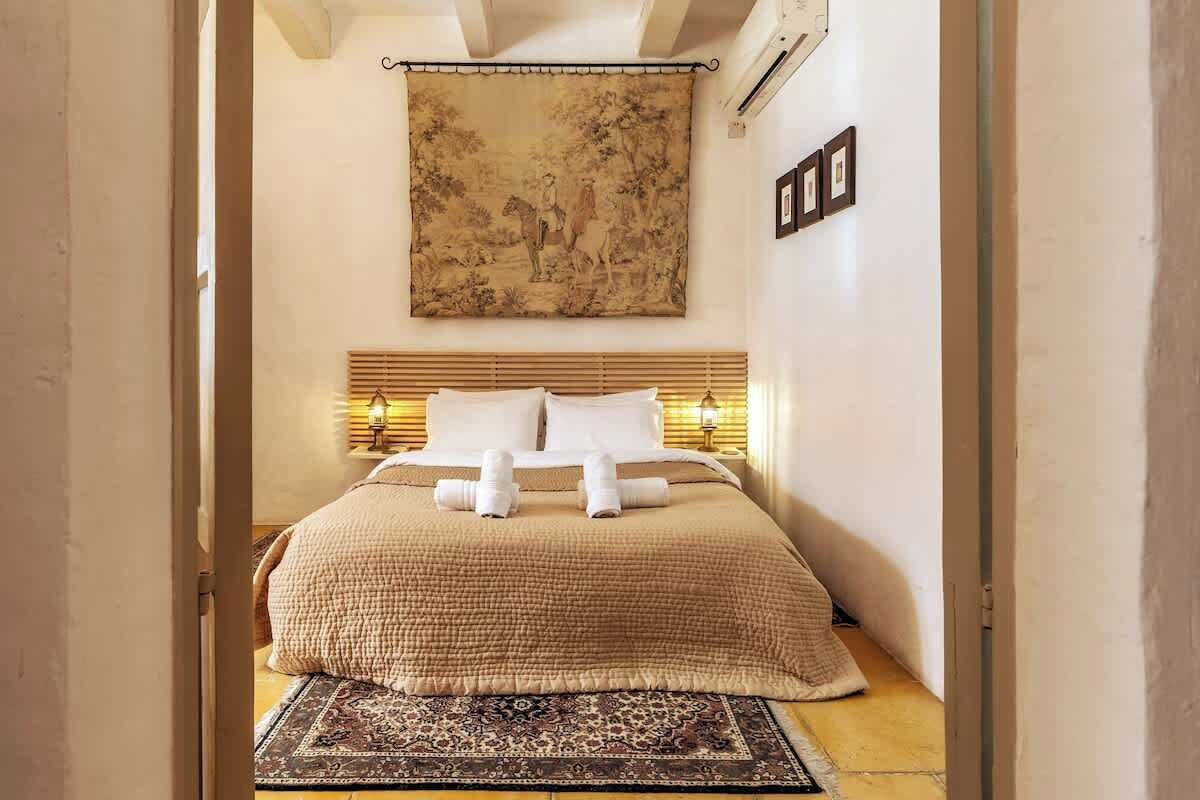 Una cama bien hecha con una colcha color beige y almohadas, un tapiz grande en la pared y una alfombra estampada en el suelo.