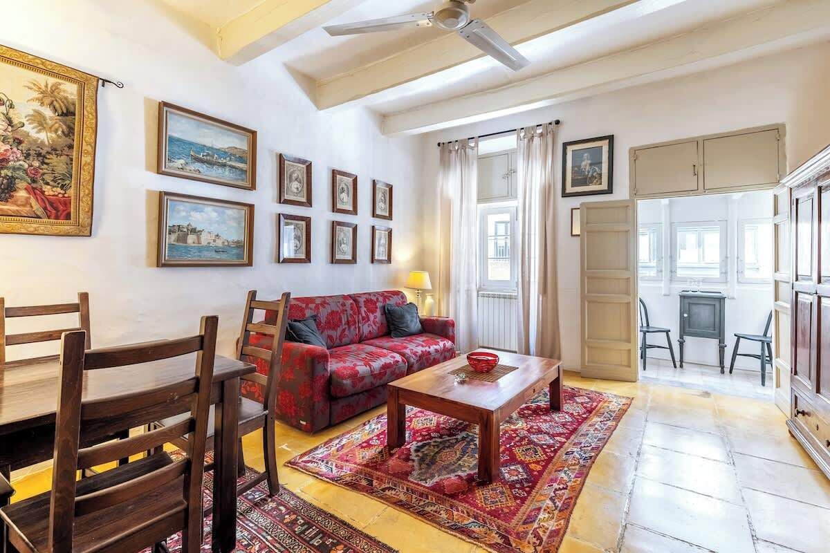 Una sala de estar con un sofá rojo, sillas de madera, una mesa de centro y una alfombra estampada. Hay cuadros enmarcados en la pared y un ventilador de techo.